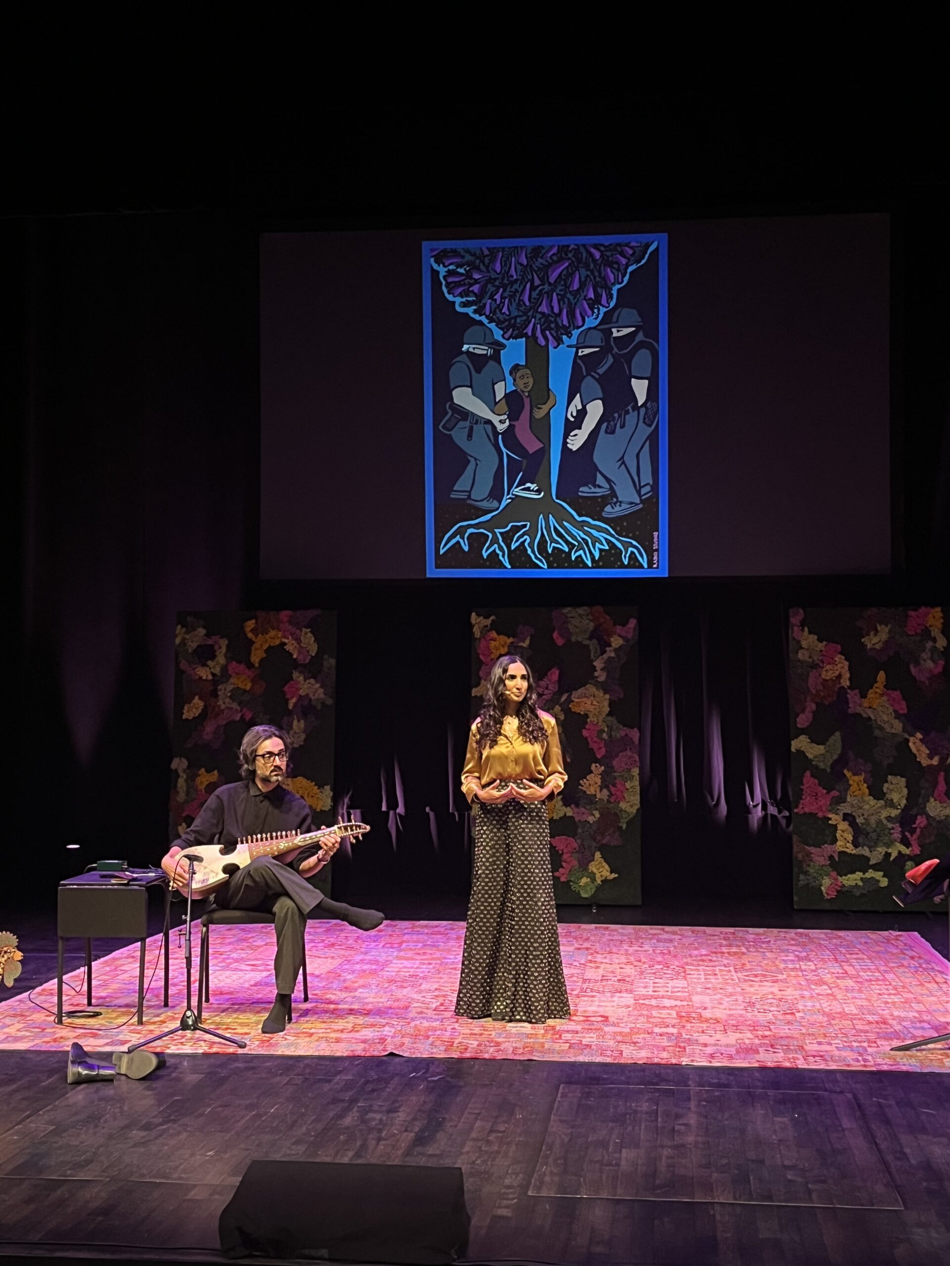 Valarie and Qais on stage at Festival of Faiths with image of jacaranda tree behind them