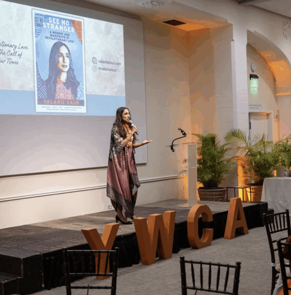 A woman stands on stage speaking to an audience in a banquet-style room. Behind her is a large screen displaying the cover of a book titled See No Stranger and her website and social media information.