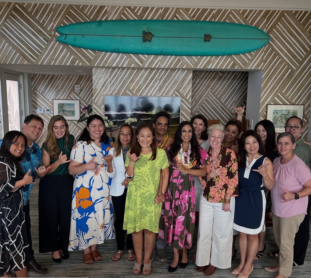 A group of people stand together indoors, smiling and making shaka hand signs. They are dressed in colorful outfits, and a long teal surfboard hangs on the wall behind them.