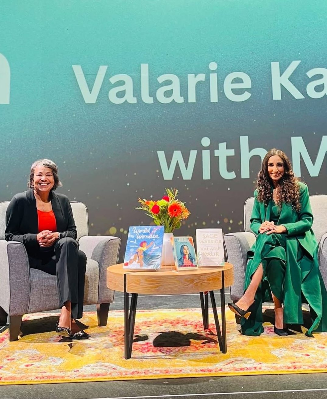 Two women sit on stage in armchairs, smiling, with a small table between them holding flowers and two books. A screen behind them displays the name “Valarie Kaur.”